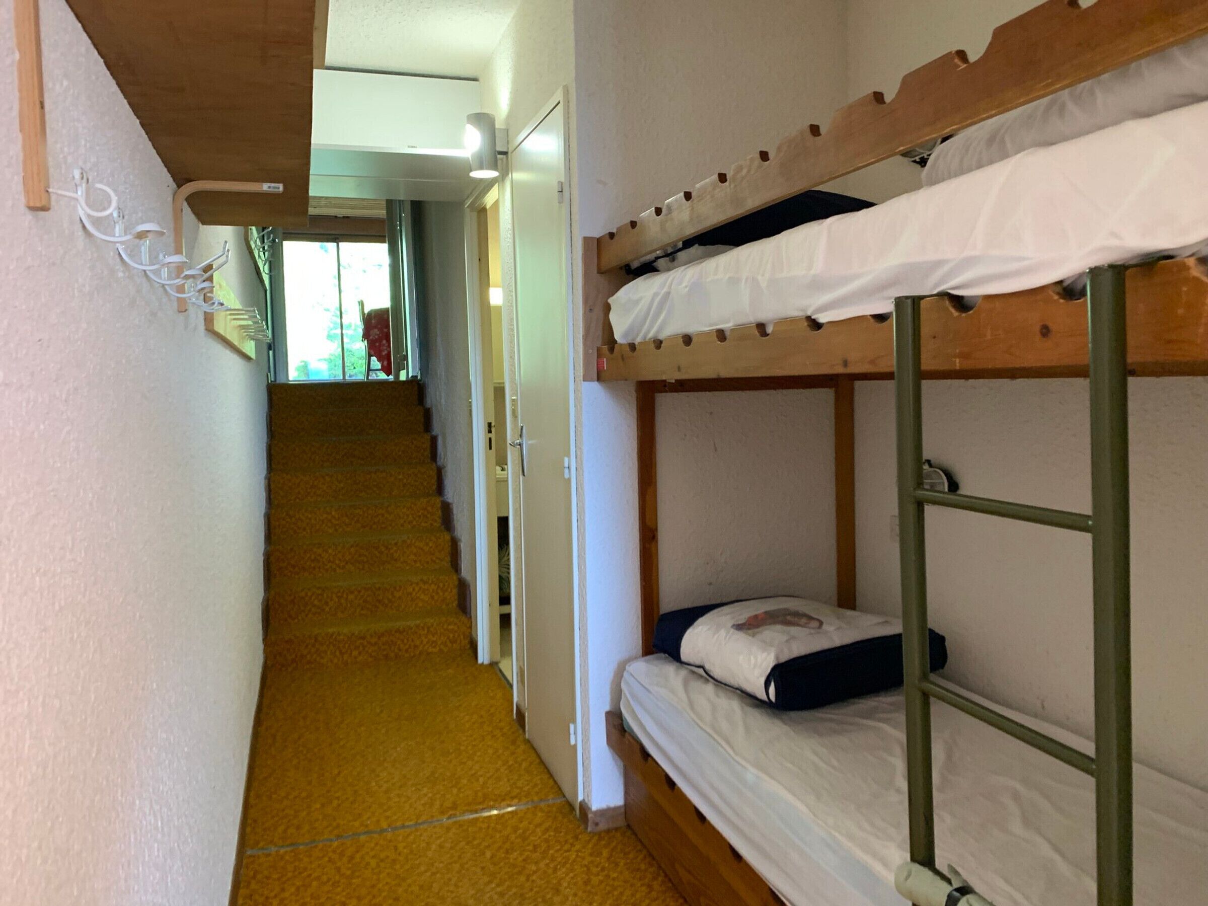 Een smalle gang met stapelbedden aan één kant en een deur aan het einde die naar een kamer leidt.