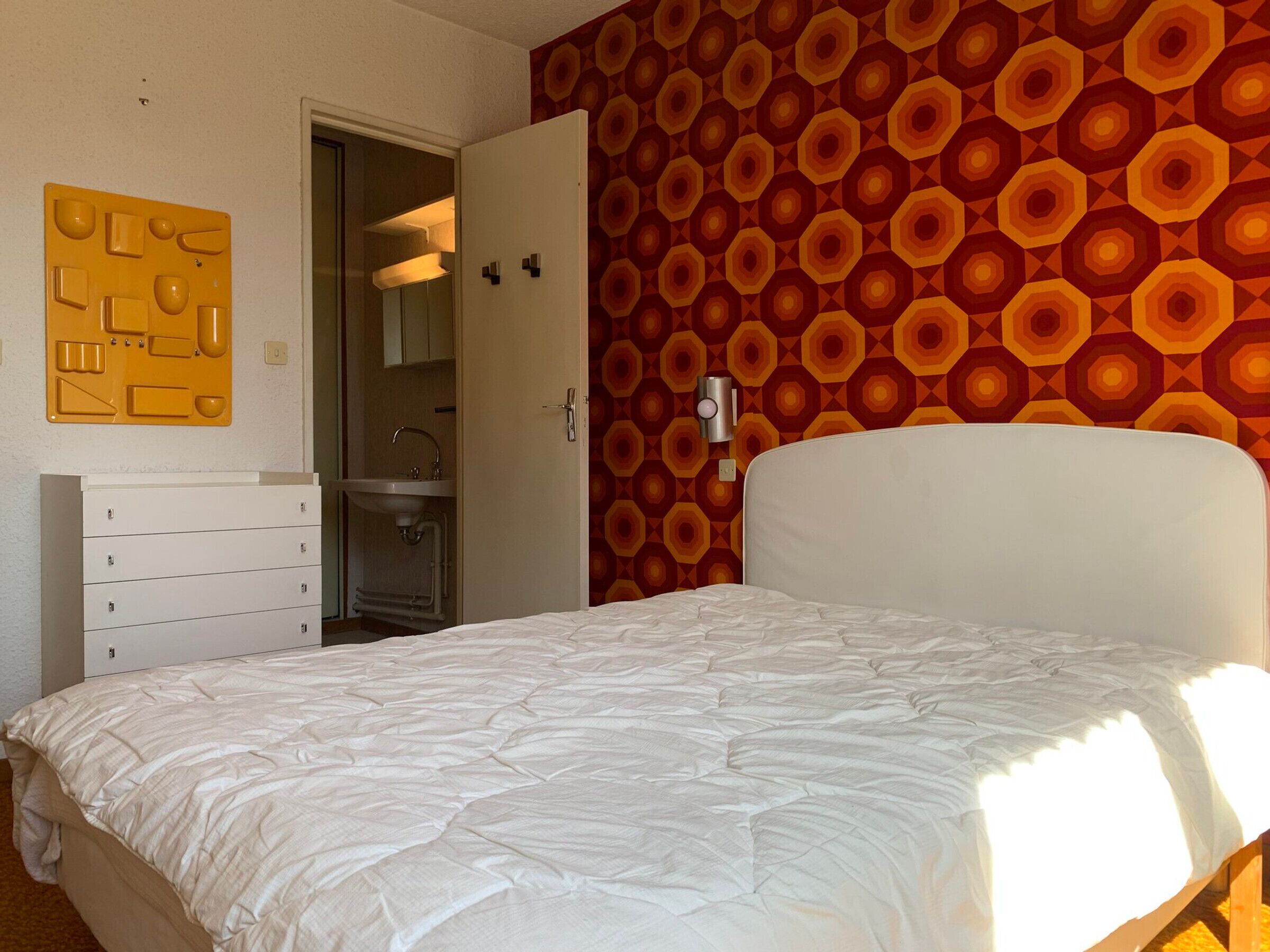 A bedroom with a bed, a white dresser, a bathroom visible through an open door, and a wall with a geometric pattern.