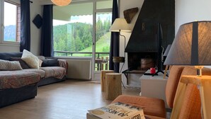 Un salon chaleureux avec un canapé, une cheminée, une lampe et une vue sur une colline boisée.
