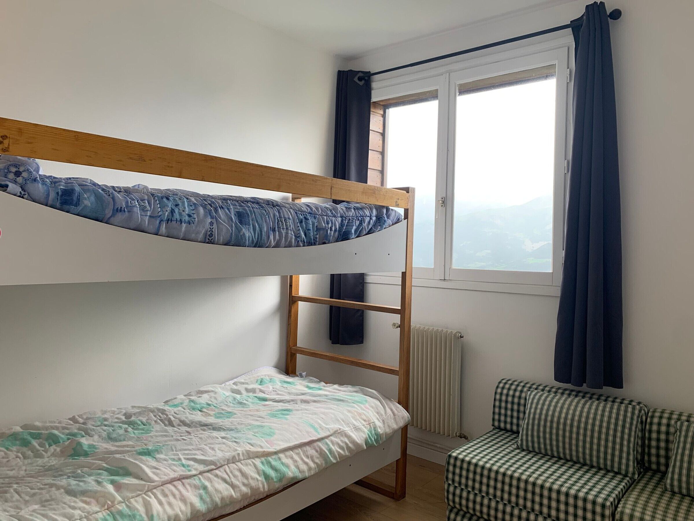 A bunk bed with a ladder, a single bed, a checkered chair, and a window with curtains.