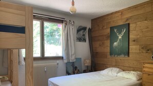 Makuuhuoneessa on kerrossänky, yksi erillinen sänky, patteri, ikkuna, jossa on verhot, lamppu ja seinällä oleva peuranaiheinen taulu.