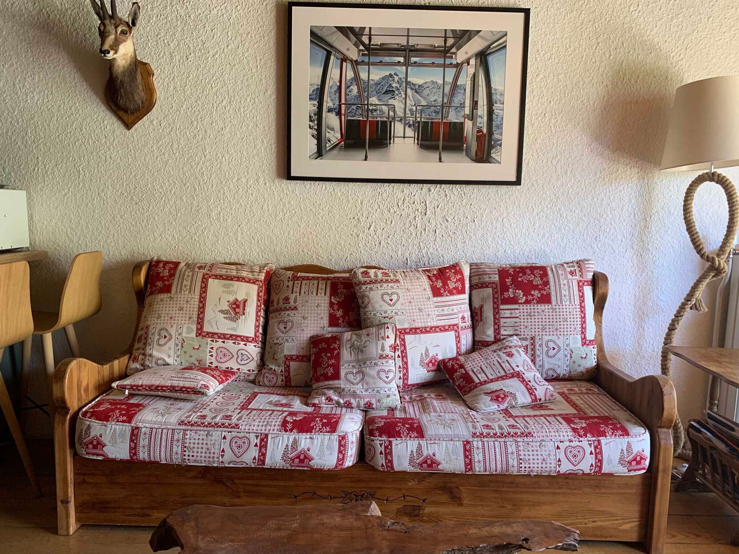 Une pièce chaleureuse avec un canapé en bois agrémenté de coussins aux motifs rouges et blancs, une tête de cerf fixée au mur et un tableau représentant un paysage de montagne.
