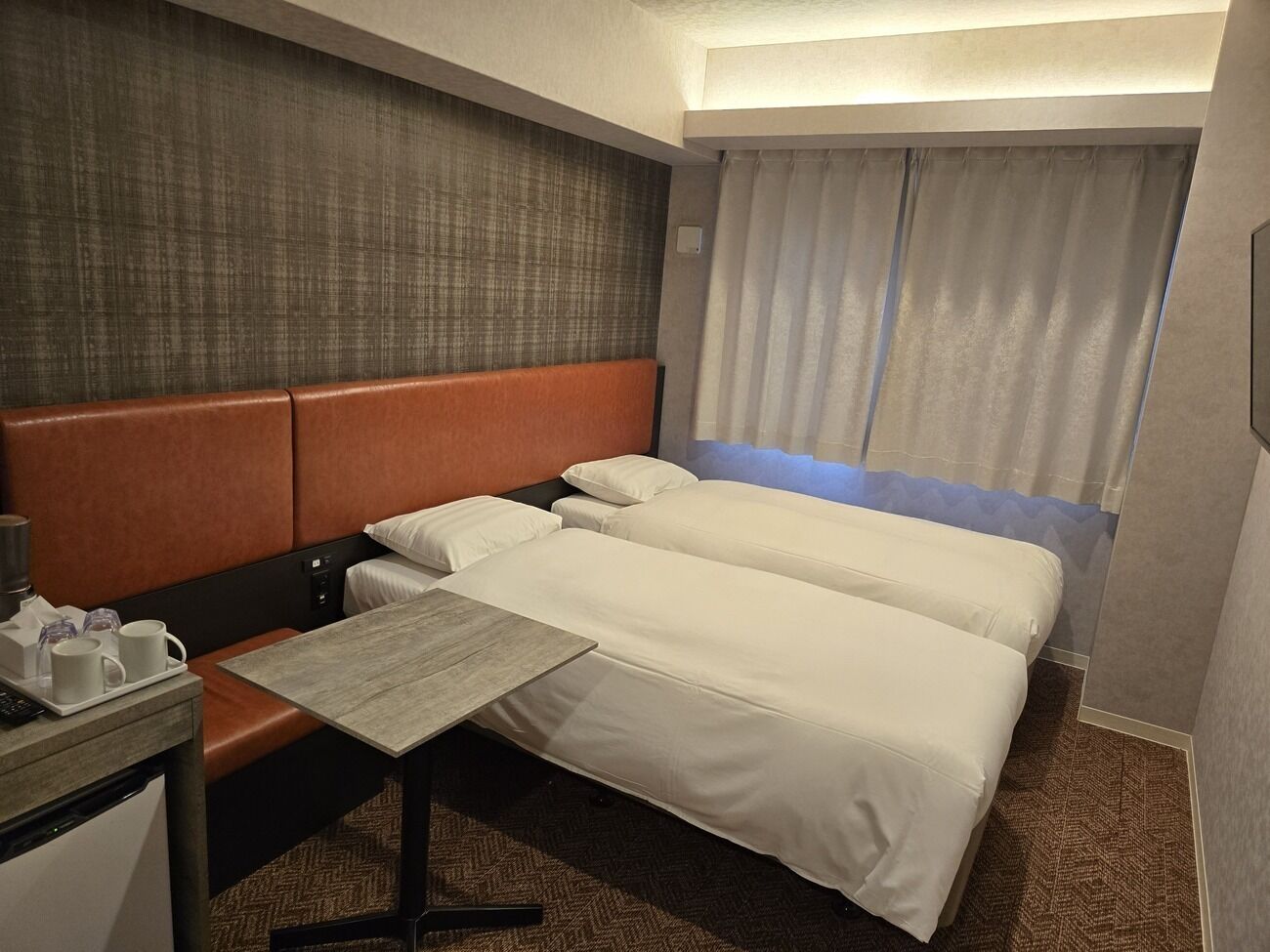 Habitación de hotel con dos camas, una mesita, un escritorio con lámpara y una ventana con cortinas.