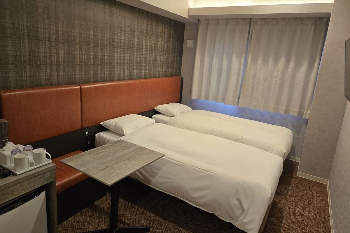Habitación de hotel con dos camas, una mesita, un escritorio con lámpara y una ventana con cortinas.
