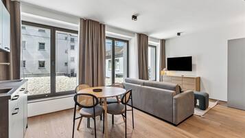 Ein modernes Wohnzimmer mit einer Couch, Esstisch und Stühlen, großen Fenstern und einem Flachbildfernseher.