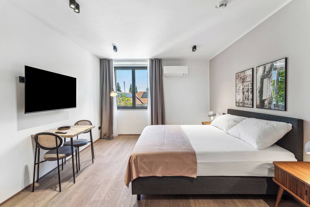 A modern hotel room with a bed, a desk with chairs, a TV, and large windows with curtains.