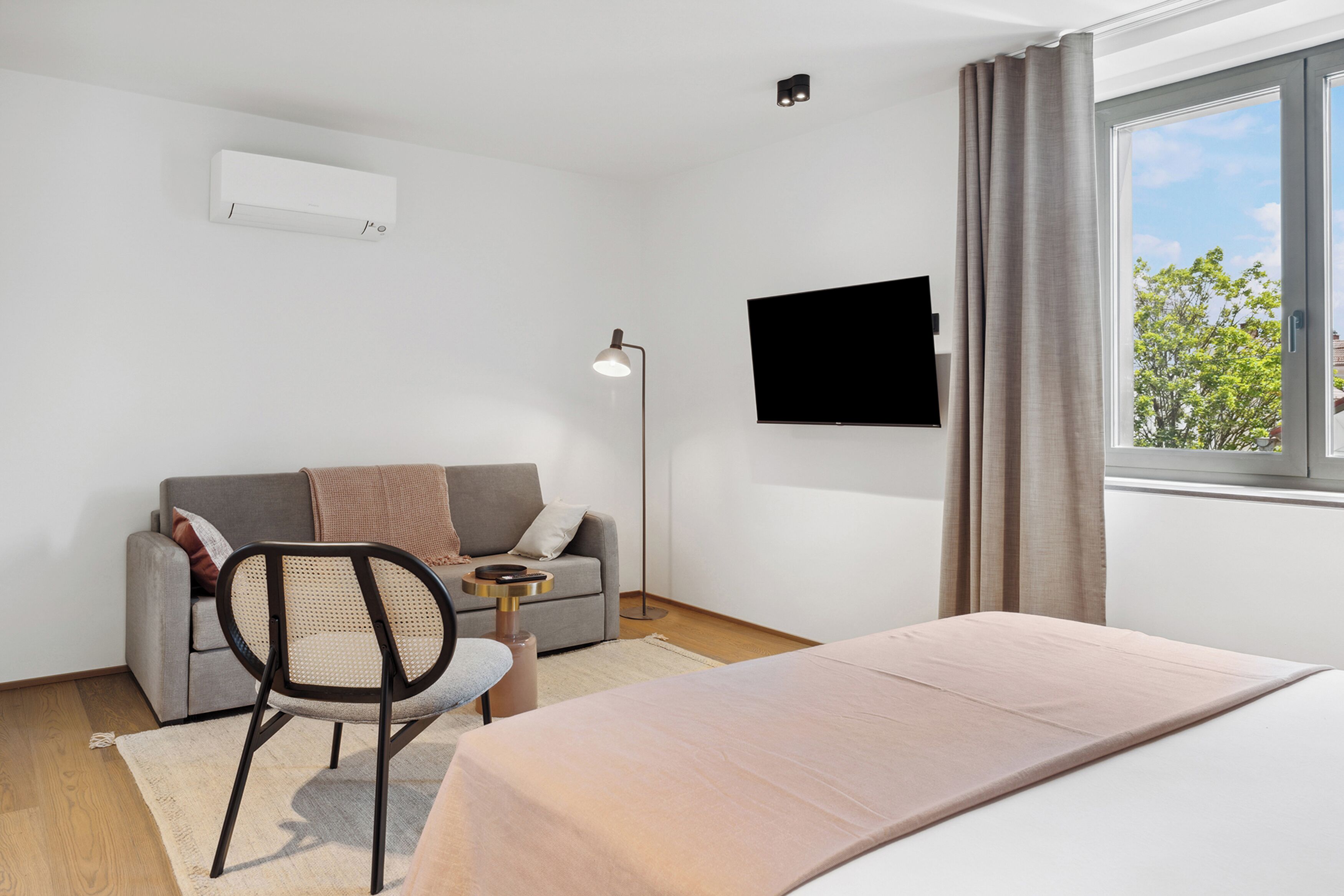 A modern hotel room with a bed, a sofa, a chair, a TV, and a window with curtains.