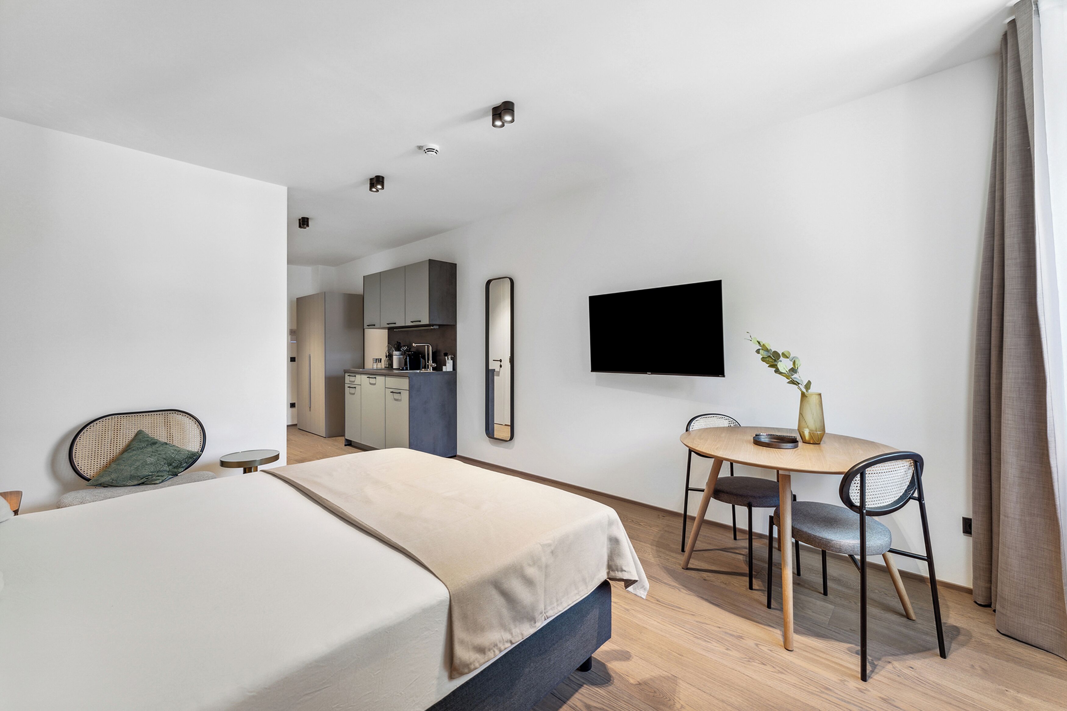 Ett modernt hotellrum med en säng, ett matbord med stolar, en tv som är monterad på väggen och ett köksutrymme i bakgrunden.