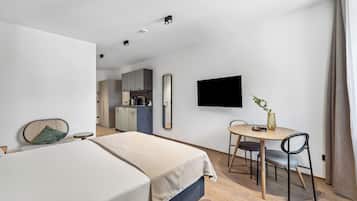 Ett modernt hotellrum med en säng, ett matbord med stolar, en tv som är monterad på väggen och ett köksutrymme i bakgrunden.