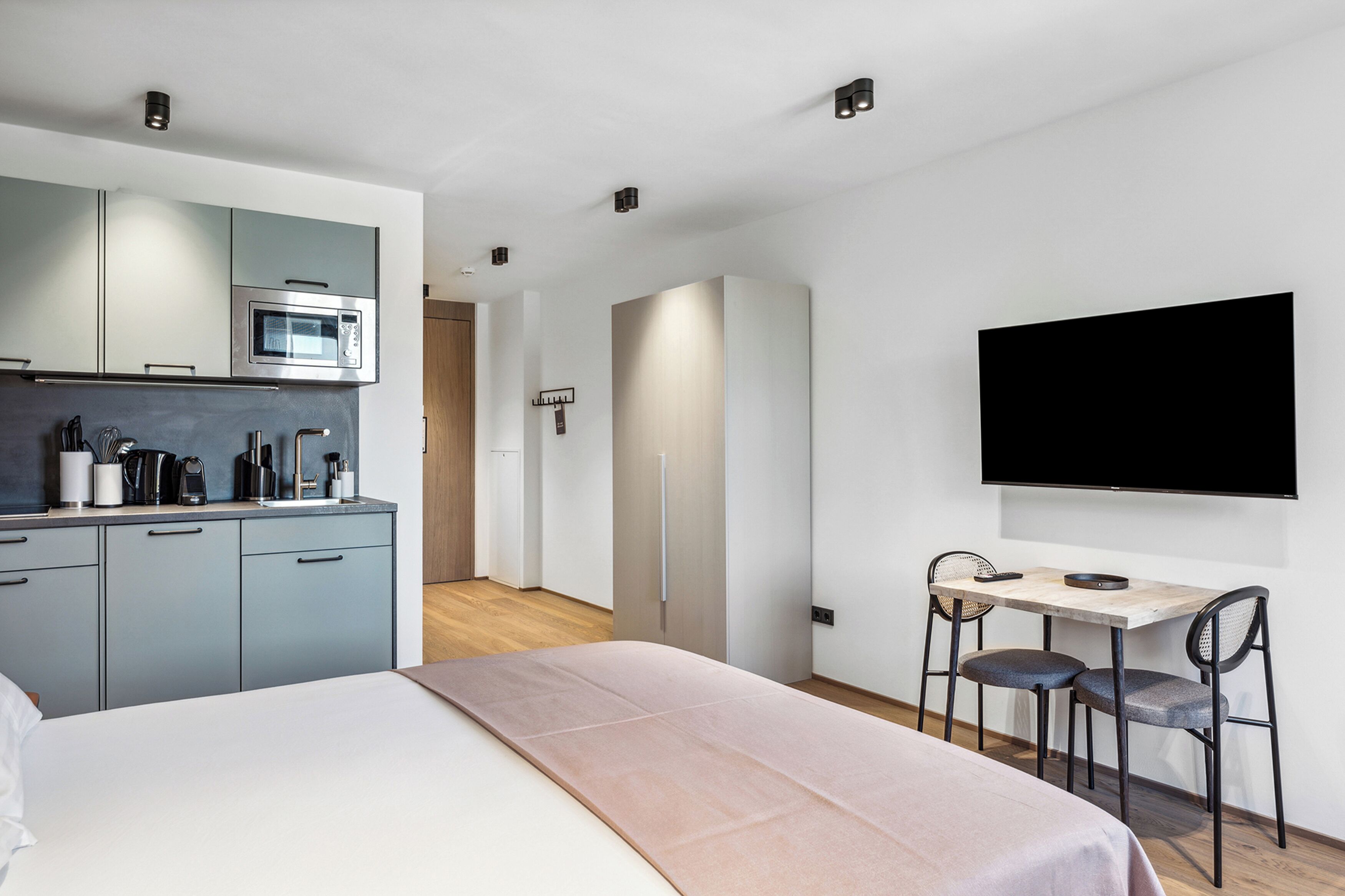 Una habitación de hotel moderna con kitchenette, una cama, un escritorio con dos sillas y un televisor montado en la pared.