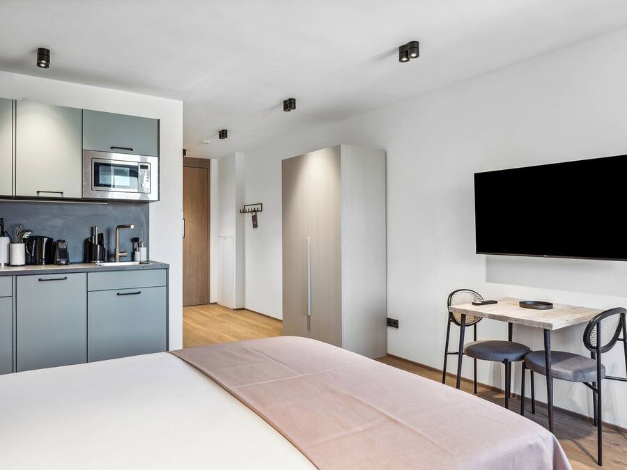 Una habitación de hotel moderna con kitchenette, una cama, un escritorio con dos sillas y un televisor montado en la pared.