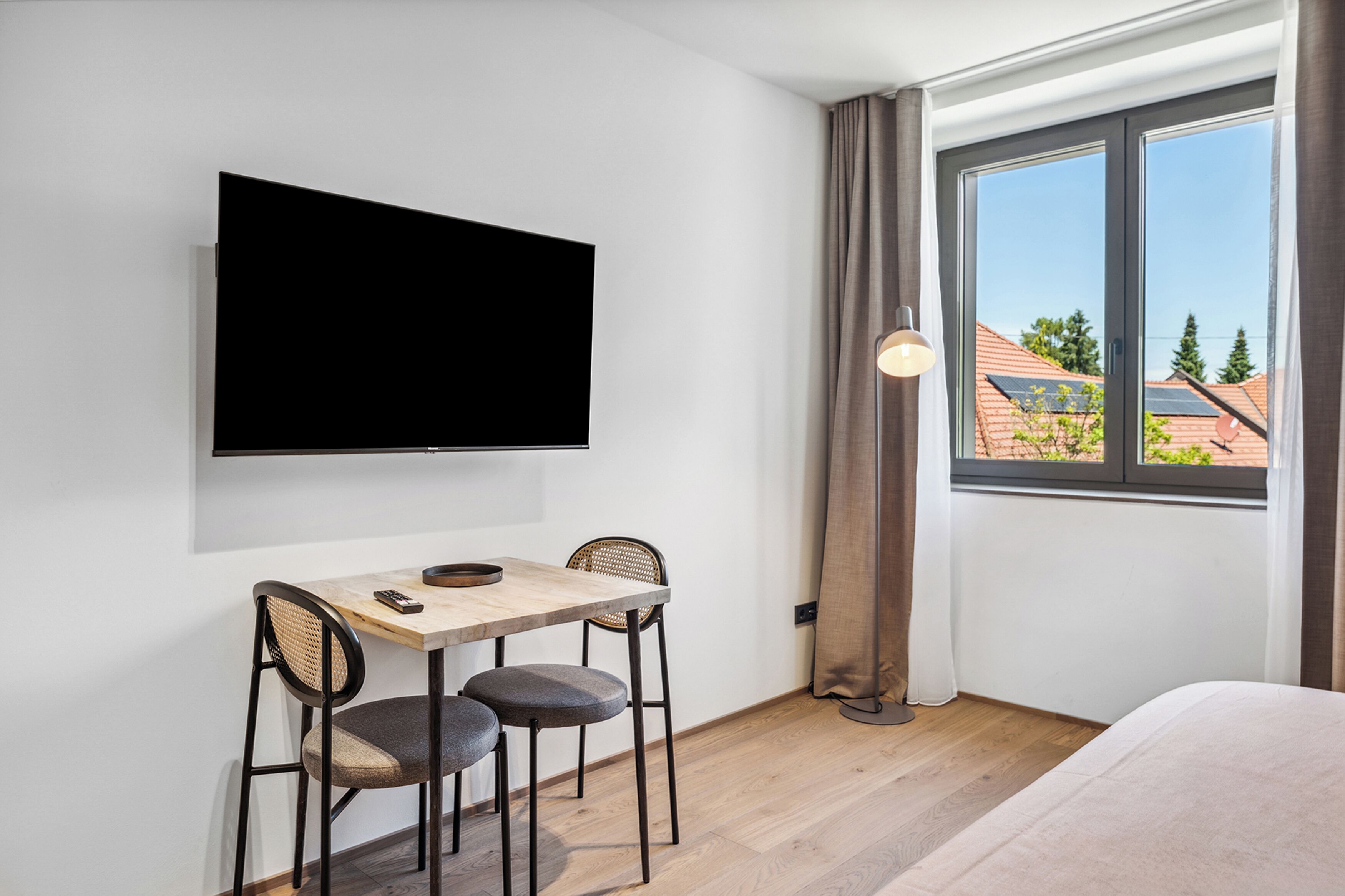 Habitación de hotel con televisión de pantalla plana montada en la pared, una mesa de madera con dos sillas, una cama y una ventana con cortinas.