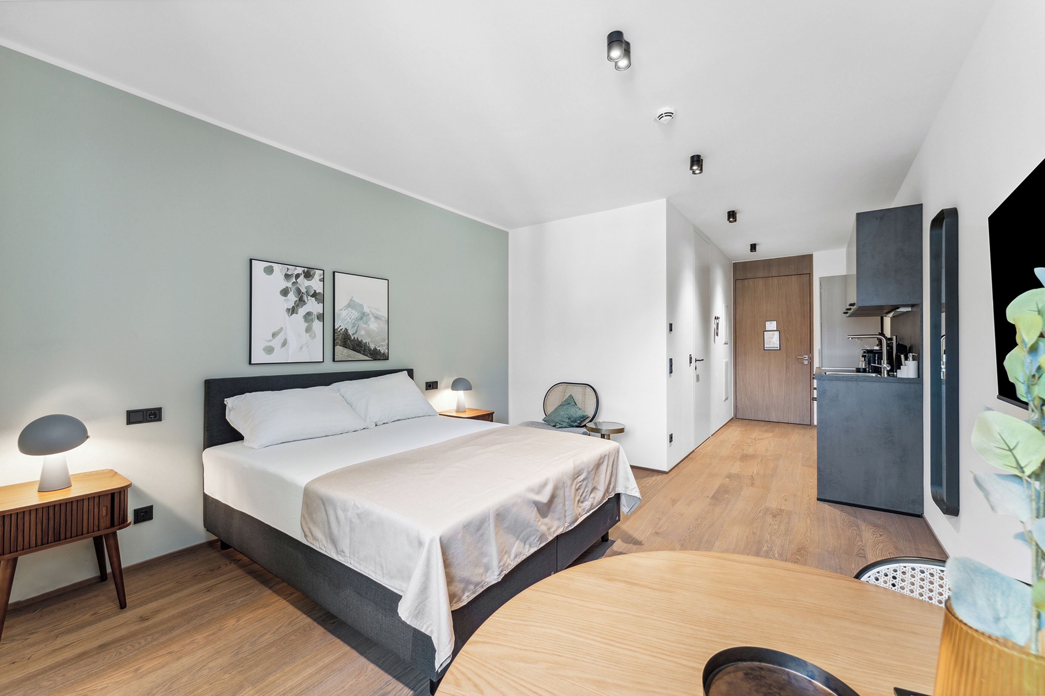 A modern bedroom with a bed, bedside tables, a desk, and a kitchenette.