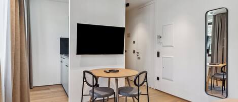 A modern hotel room with a flat-screen TV mounted on the wall, a round wooden table with two chairs, a bed with a headboard, and a large mirror on the wall.
