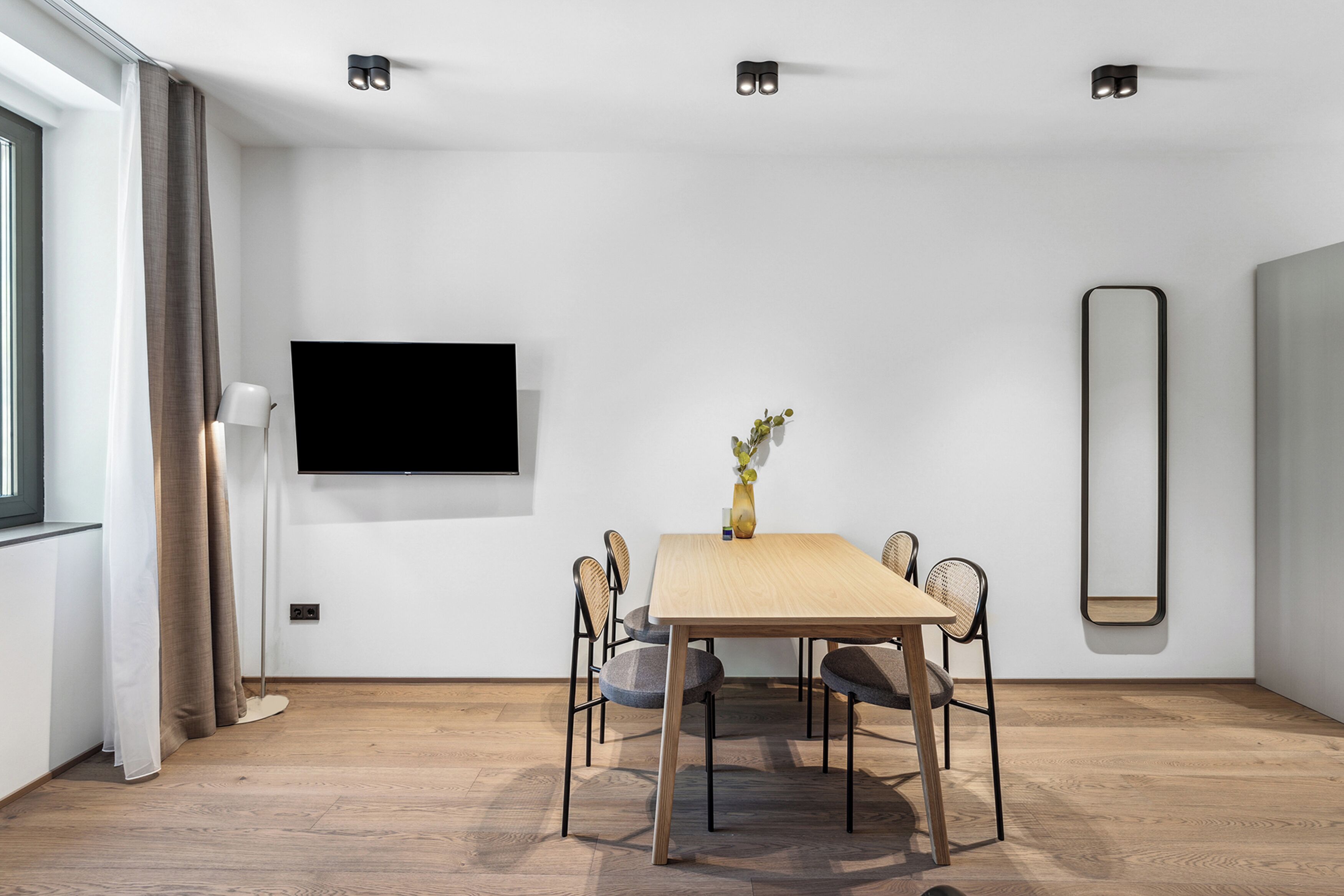 Un'elegante zona pranzo moderna con tavolo in legno, quattro sedie, un vaso con fiori, una TV a schermo piatto e un grande specchio a parete.