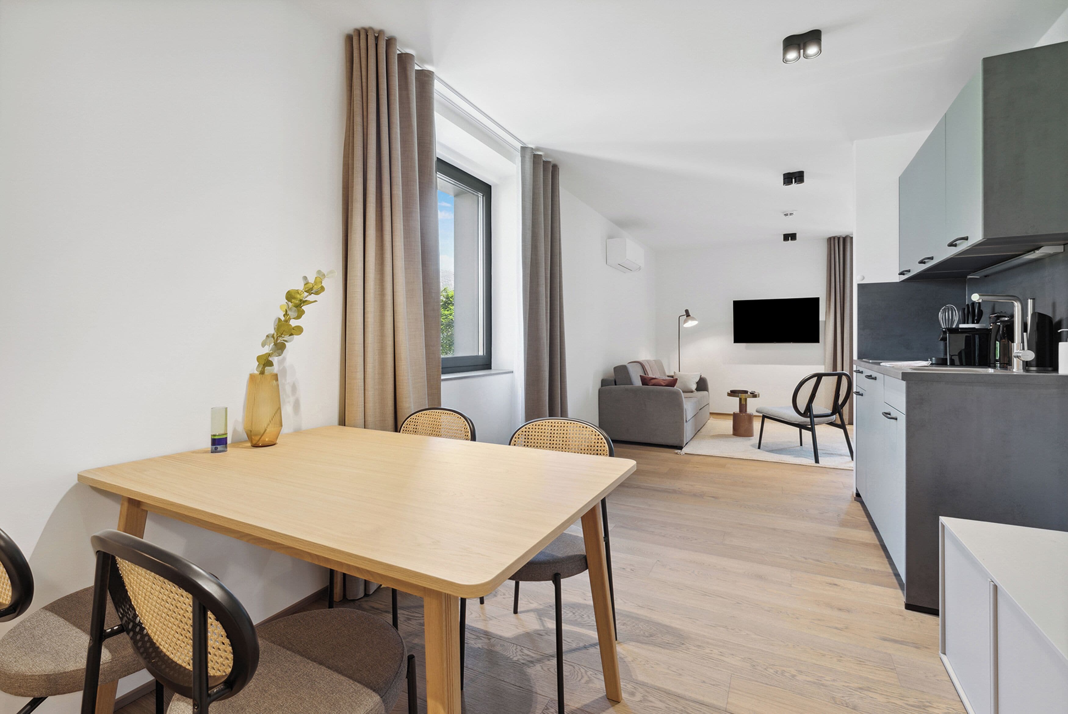 Ett modernt kök med en platt-TV, en matplats med ett träbord och stolar samt ett vardagsrum med en soffa och ett litet sidobord.