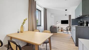 Ett modernt kök med en platt-TV, en matplats med ett träbord och stolar samt ett vardagsrum med en soffa och ett litet sidobord.