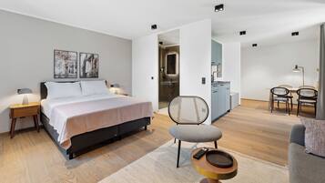 A modern bedroom with a bed, bedside tables, a chair, a small table, and a bathroom visible through an open door.
