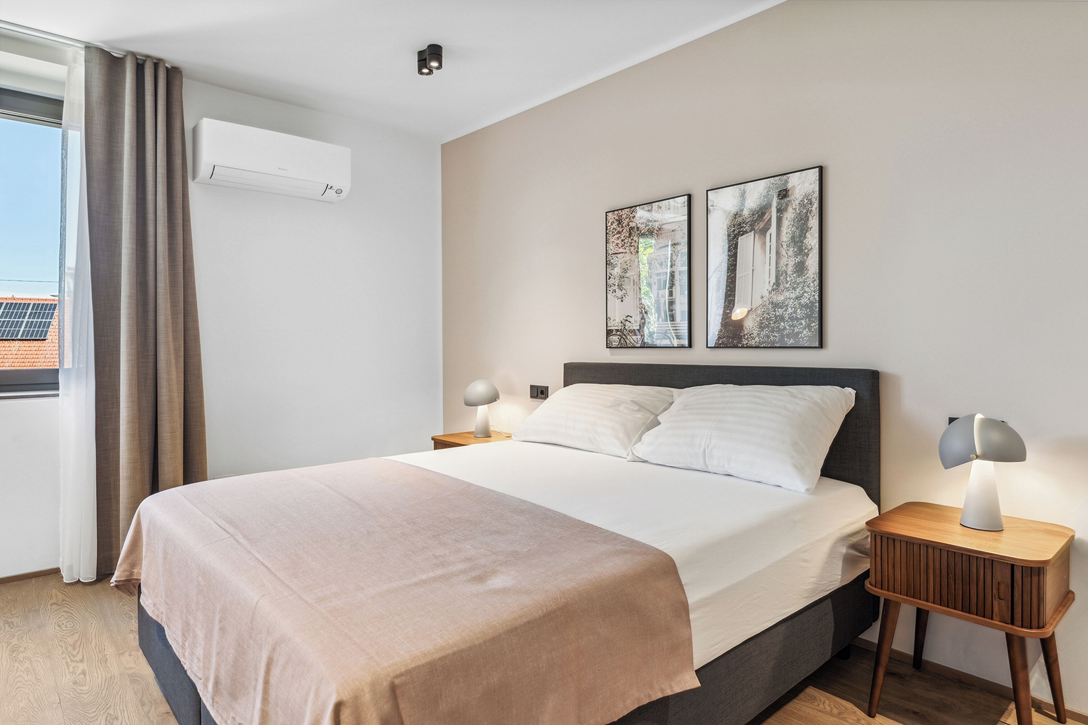 Una habitación de hotel moderna con una cama grande, dos mesitas de noche con lámparas, una ventana con cortinas y dos obras de arte colgadas en la pared.