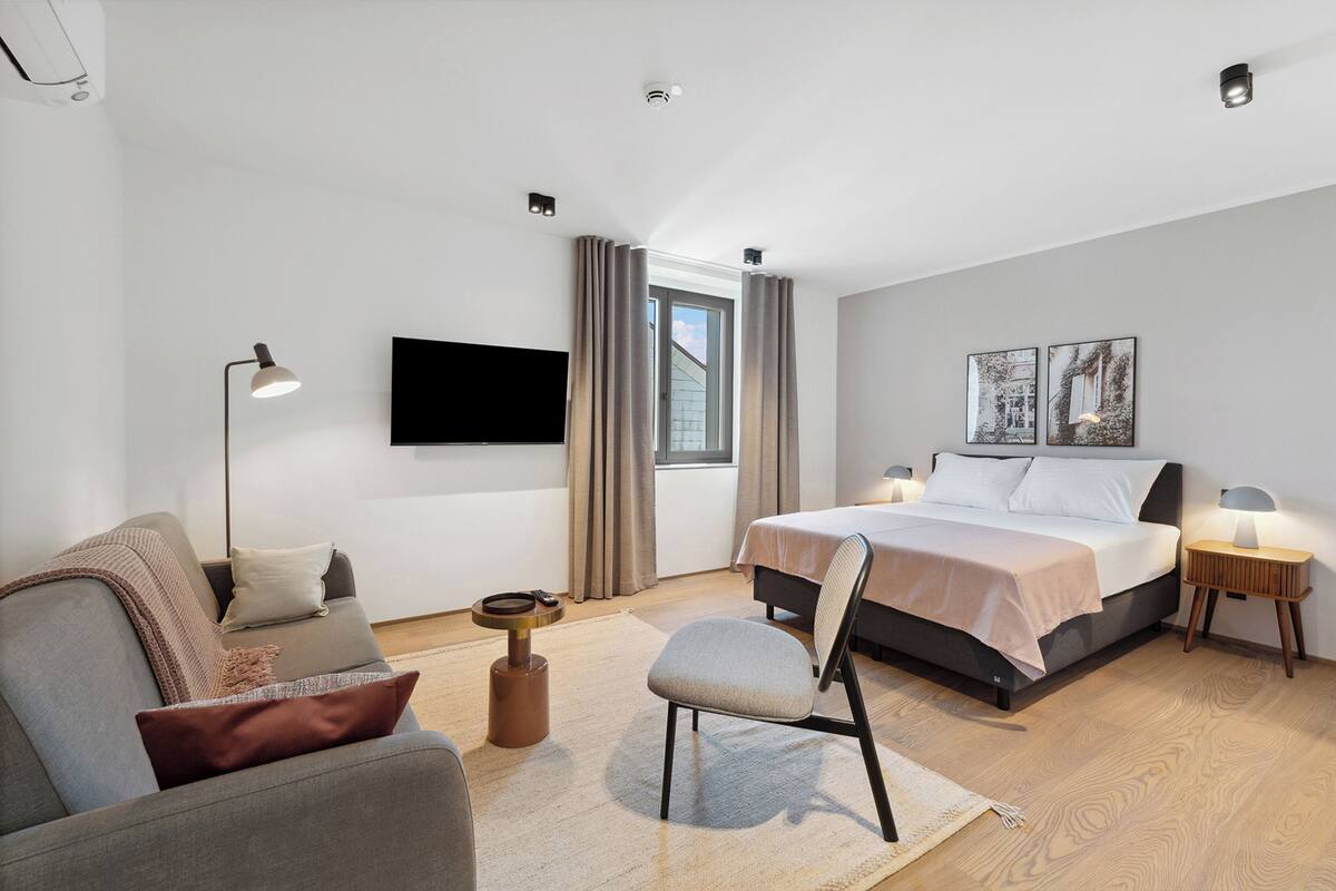 A modern hotel room with a bed, a sofa, a chair, a small table, a TV, and a window with curtains.