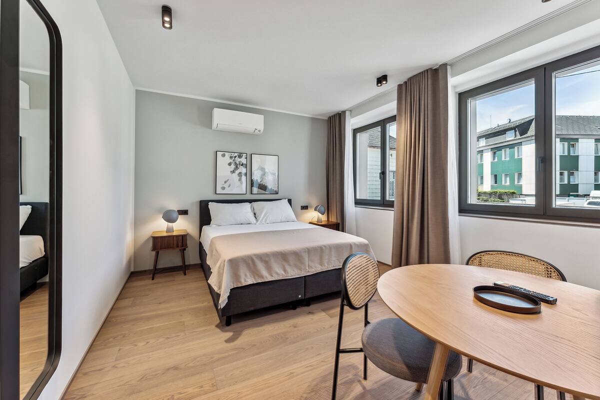 A hotel room with a bed, bedside table, lamp, and a view of buildings through the window.
