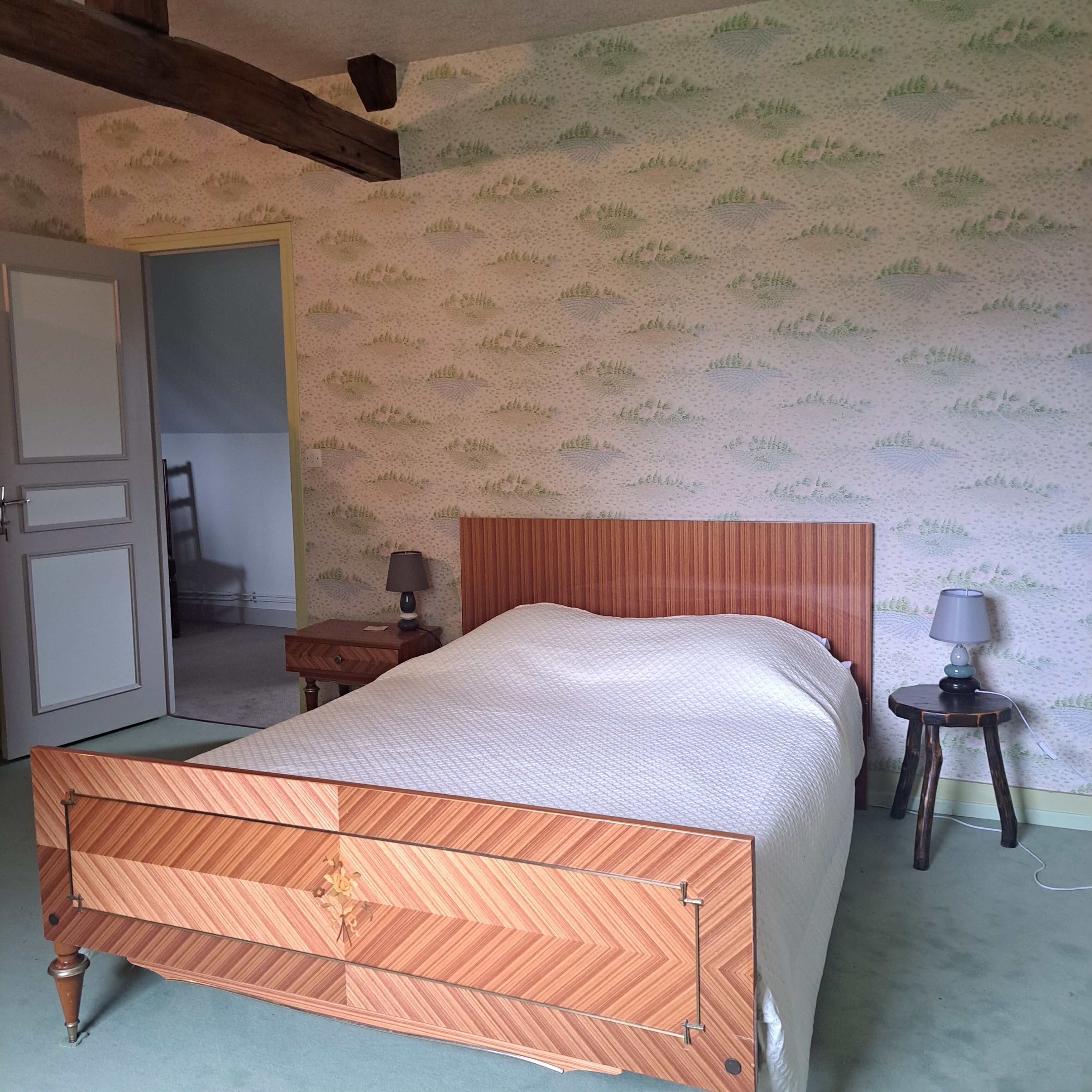 A bedroom with a wooden bed, a nightstand, a lamp, and a door leading to another room.