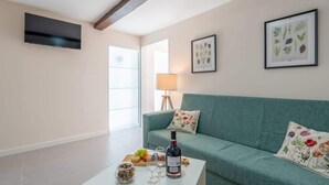 A living room with a teal sofa, a coffee table with a bottle, glasses, and snacks, a TV mounted on the wall, and framed botanical prints.