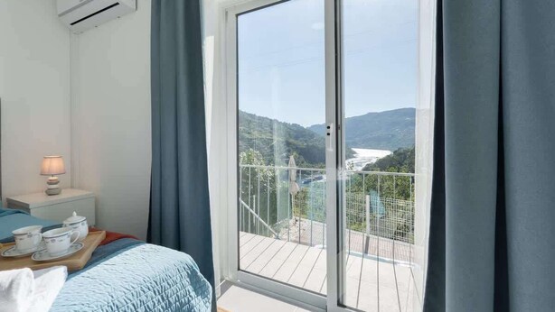 A bedroom with a bed, a nightstand, a tray with cups and saucers, and a view of a balcony with a railing and greenery.