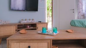 Ein Zimmer mit Holztisch, Korbmöbeln, einem Flachbildfernseher und Blick auf Grünflächen.