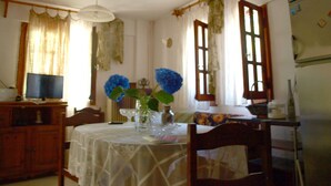 A dining area with a table covered in a white tablecloth, chairs, a vase with blue flowers, a television on a wooden cabinet, and a refrigerator in the background. - Paradeisos Studio With Garden View (Zagora-Mouresi)
