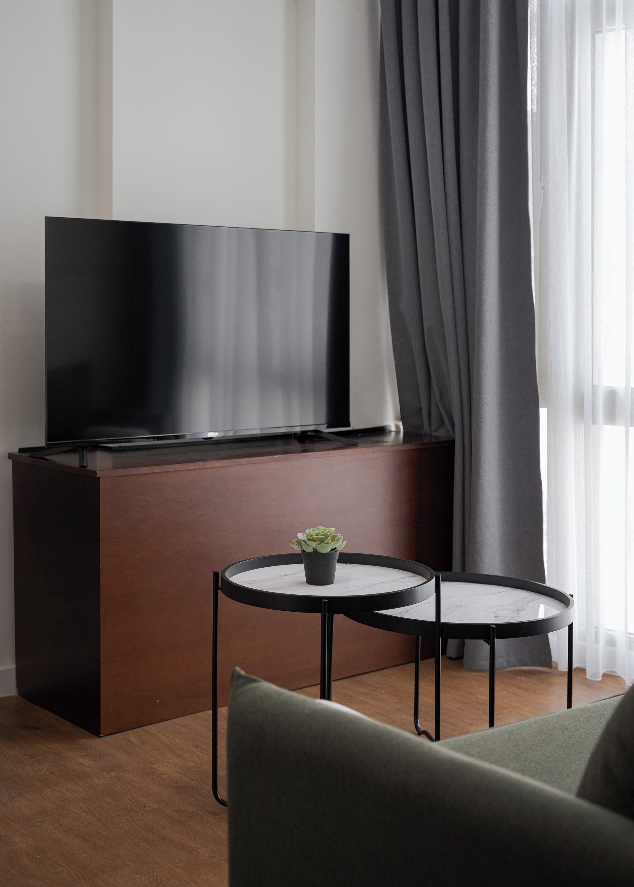 A modern living room with a flat-screen TV on a wooden cabinet, two round tables with marble tops, a small potted plant, and a grey sofa.