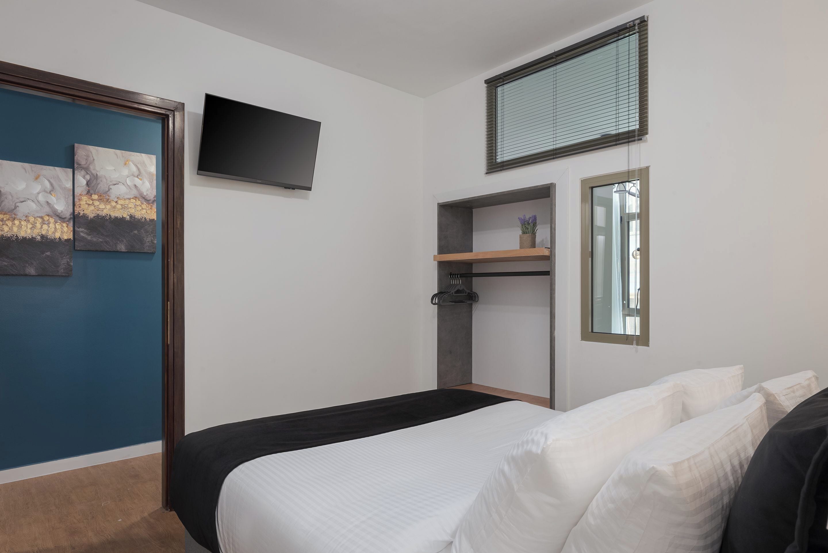 Une chambre à coucher avec un lit, une télévision, une fenêtre avec des stores, une étagère avec une plante et des œuvres d’art accrochées au mur.