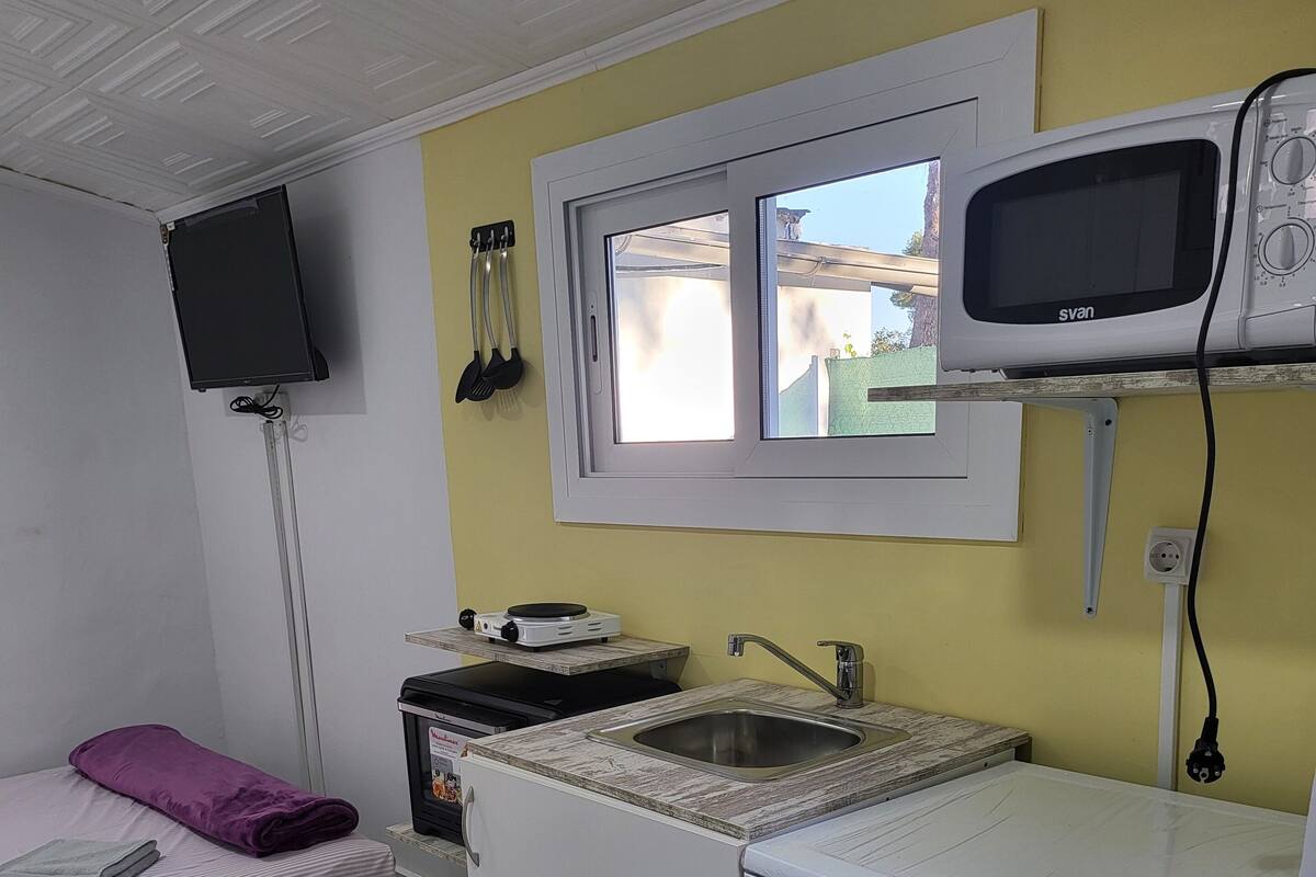 A compact kitchen with a microwave, sink, and a small refrigerator. There's a window above the sink, and a television mounted on the wall.
