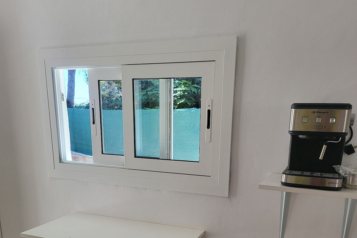 A room with a white table, two stools, a window, and a coffee machine.