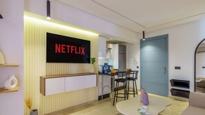 Un salon moderne avec un téléviseur à écran plat affichant le logo « NETFLIX », un coin repas avec une table et des chaises, et une cuisine en arrière-plan.