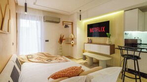 Une chambre d’hôtel moderne équipée d’un lit, d’un bureau, d’un tabouret de bar, d’un téléviseur affichant « NETFLIX » et d’une étagère murale agrémentée d’objets décoratifs.