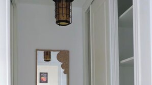 Un pasillo con una lámpara colgante, un espejo y una puerta que da a una habitación con otra puerta blanca y un estante.