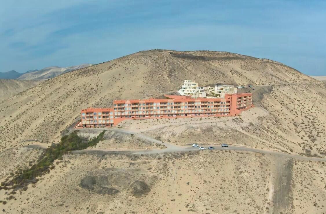 Un hotel de varios pisos con fachada roja, ubicado en una ladera con paisaje desértico al fondo.