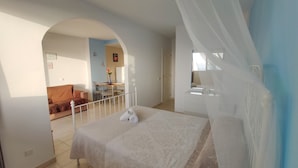 Un dormitori amb llit, finestra amb cortines transparents, balcó i zona de dinar al fons.