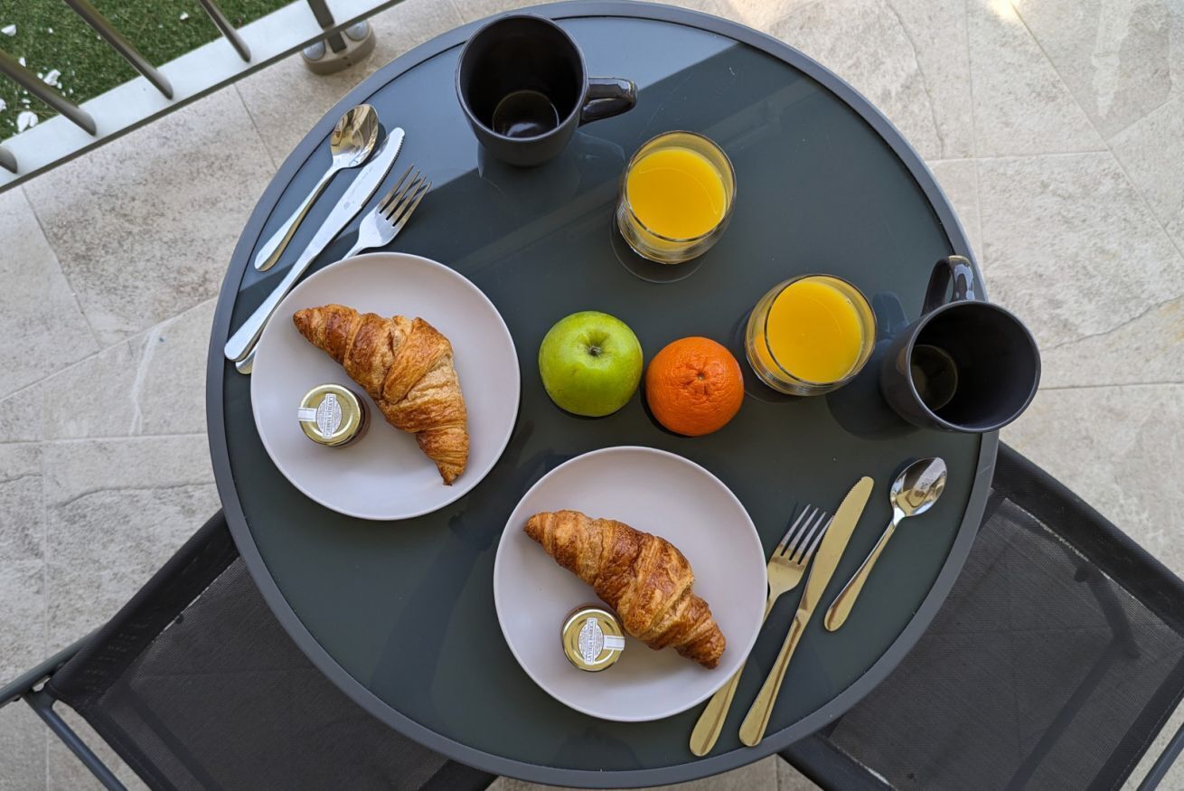A round glass table with breakfast items including croissants, an apple, an orange, and two glasses of orange juice.