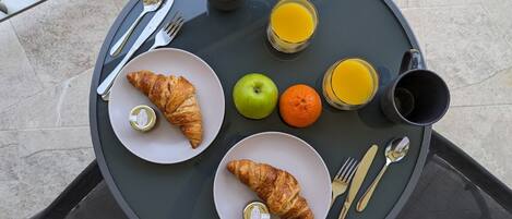 A round glass table with breakfast items including croissants, an apple, an orange, and two glasses of orange juice.