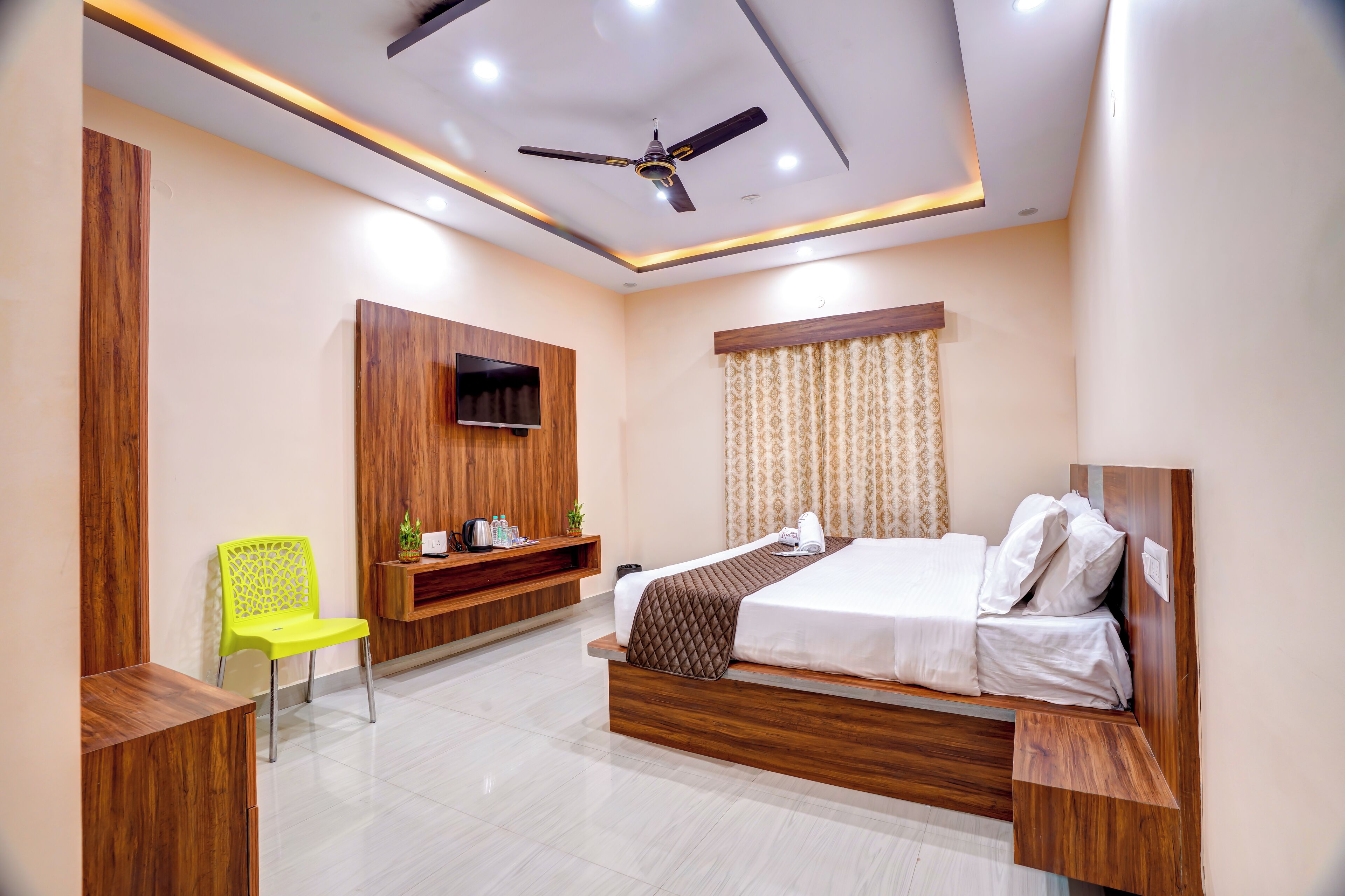 Un dormitorio moderno con una cama, un televisor montado en la pared, un ventilador de techo y una silla amarillo brillante.