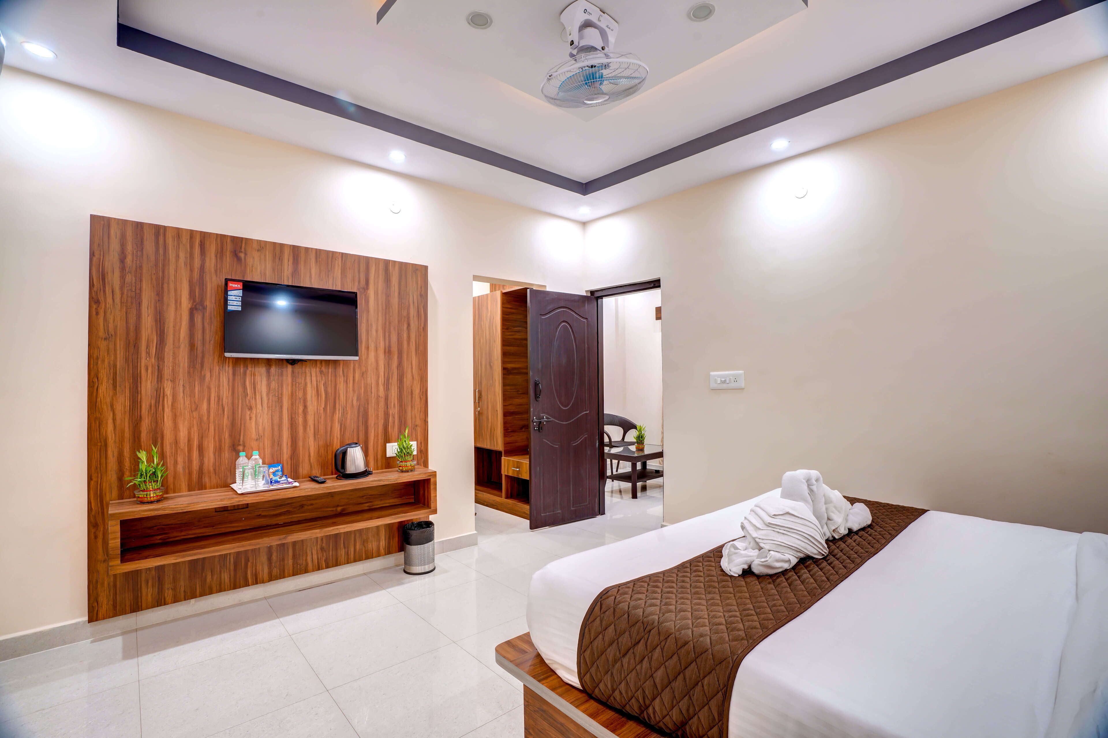 A modern hotel room with a large bed, a TV mounted on a wooden panel, a small desk with a chair, and a door leading to a dining area.