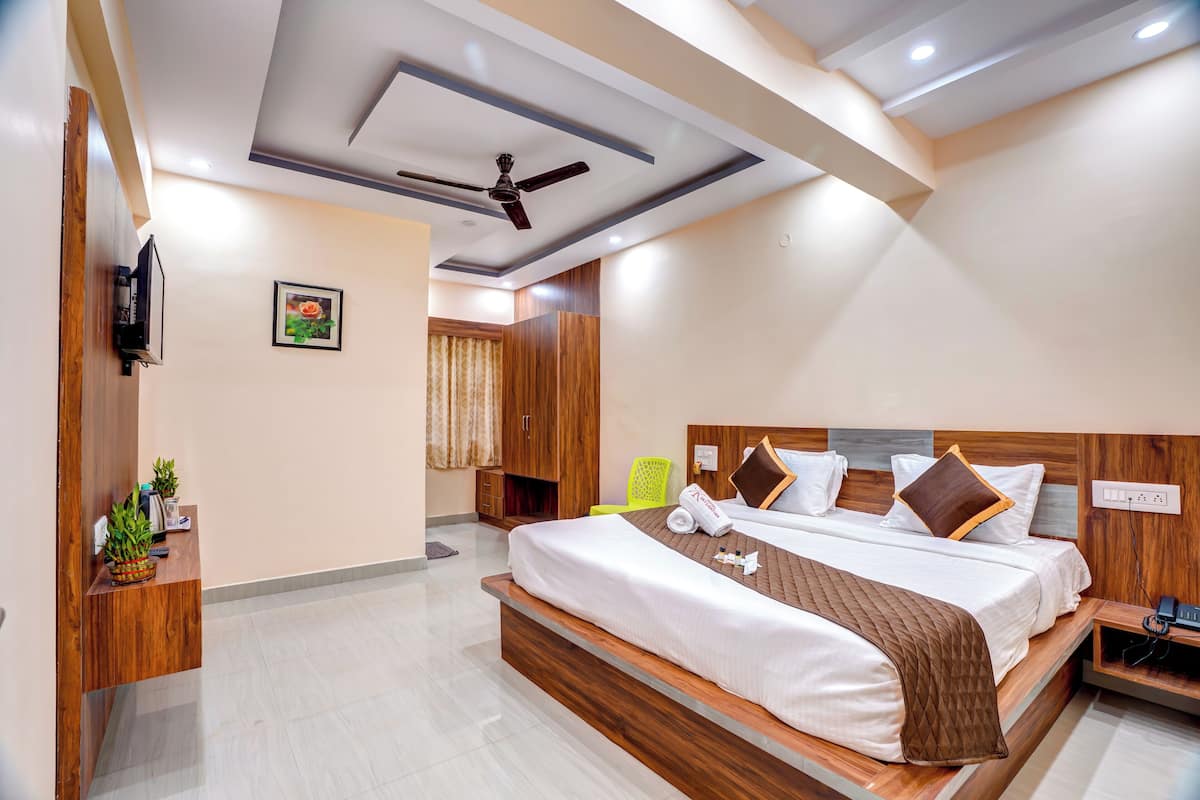 A modern hotel room with a large bed, wooden headboard, a ceiling fan, a TV, and a reception desk with plants.
