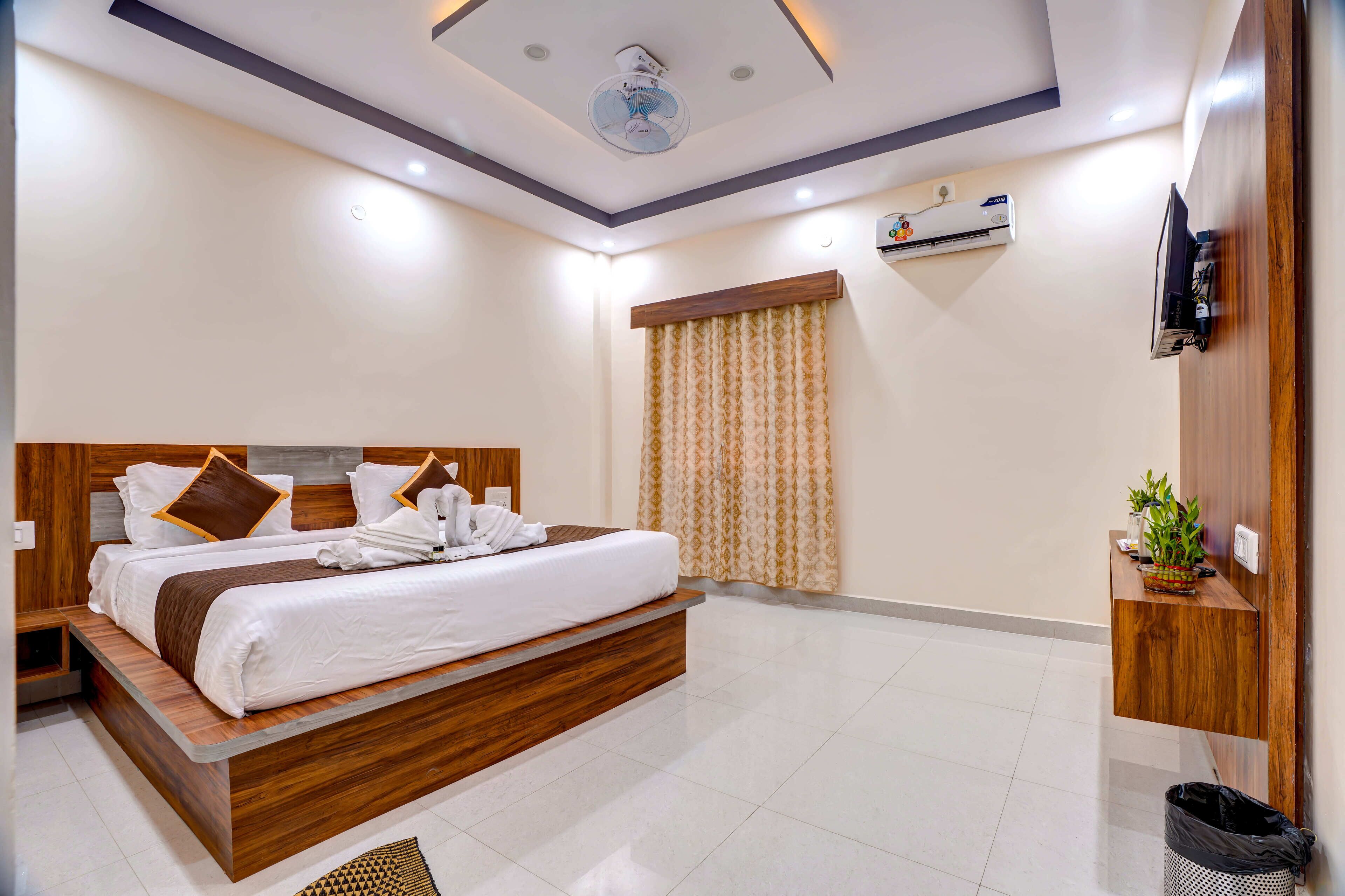 A modern hotel room with a wooden bed, a ceiling fan, an air conditioning unit, a TV, and a small plant on a wooden shelf.