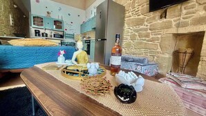 A cozy living room with a bed, a wooden coffee table with decorative items, a kitchen area with light blue cabinets, and a stone fireplace. - Rustic 2 Bedroom Stone Apartment in Malonas Rodos (Malonas Rodos)