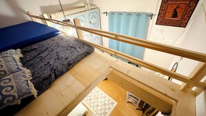 Une chambre de type dortoir, avec un lit superposé, une petite fenêtre avec un rideau et une étagère fixée au mur, ornée d’objets décoratifs.