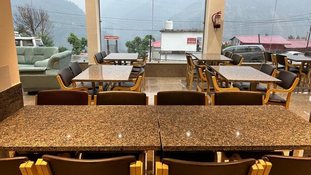 Un restaurante con mesas y sillas de granito, ventanas amplias con vistas a montañas y coches estacionados.