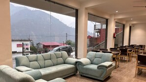 Amplia habitación con un ventanal que ofrece vistas a montañas y un edificio de tejados rojos. Hay dos sofás azul claro y una zona de comedor con mesas y sillas.