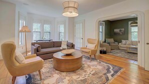 A spacious living room with a large sofa, two armchairs, a coffee table, and a patterned rug. There are large windows providing natural light and a view of greenery outside. - Short Drive to Gray's Beach! (Yarmouth)